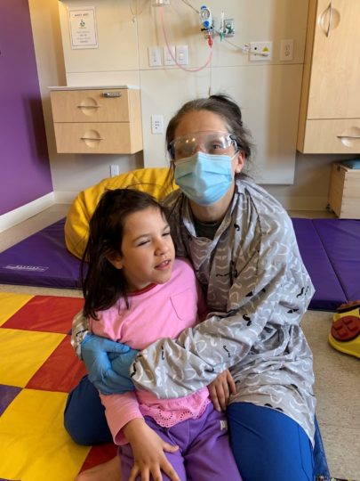 An Emily's House nurse, wearing a mask for pandemic protocols, gives a hug to a child while playing on the floor together