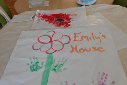 A photo of child art with flowers made of children’s hands and the painted words Emily’s House.