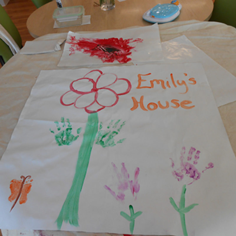 A photo of child art with flowers made of children’s hands and the painted words Emily’s House.