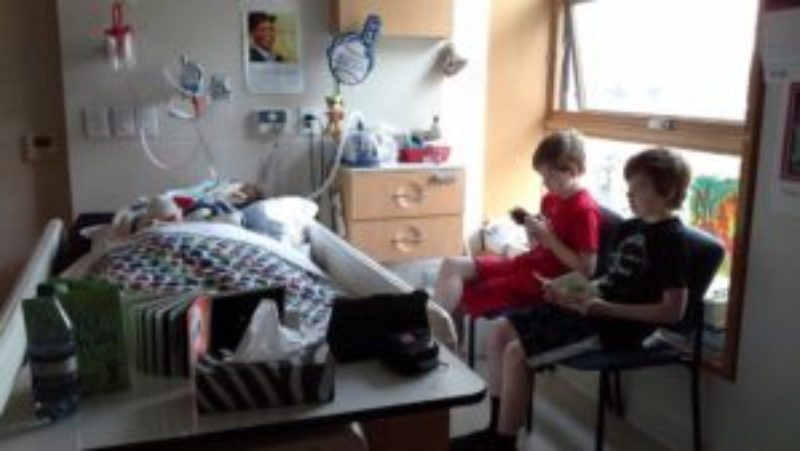 A photo of Matthew in a hospital bed at Emily’s House children’s hospice as his twin brothers sit nearby playing computer games and reading