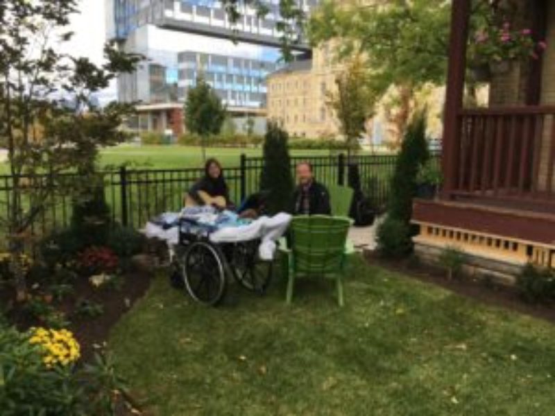 This photo of an outdoor Music Therapy session features “Luke” reclining in a wheelchair in the Emily’s House backyard garden, as his dad sits in a chair, and the music therapist plays guitar