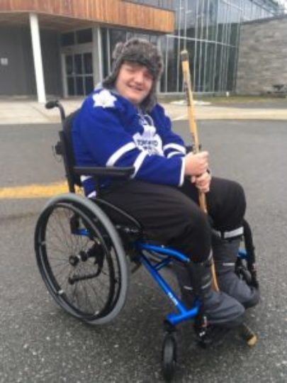 Photo of Owen wearing a Toronto Maple Leafs jersey and holding a hockey stick as he sits in a wheelchair in front of a recreation centre