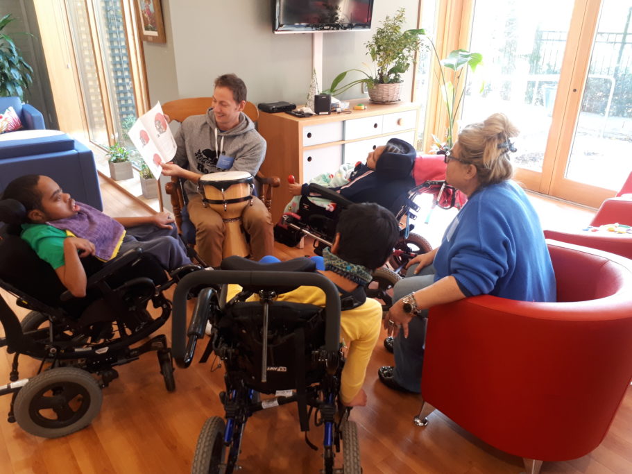 A volunteer reads a picture book to three Emily’s House kids in wheelchairs, in a sunny room.